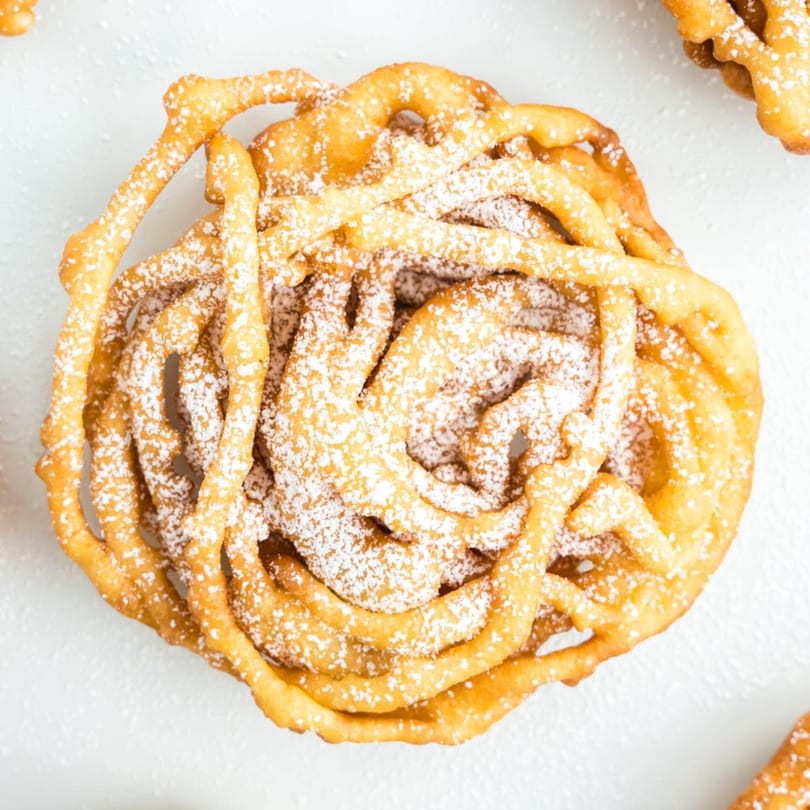 Golden-brown funnel cake, dusted with powdered sugar, on a white surface.