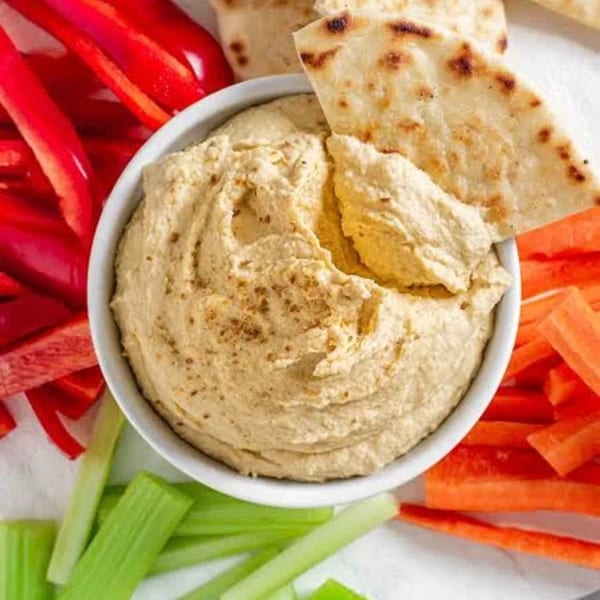 Hummus in a white bowl with pita bread, red bell pepper, carrots, and celery sticks.
