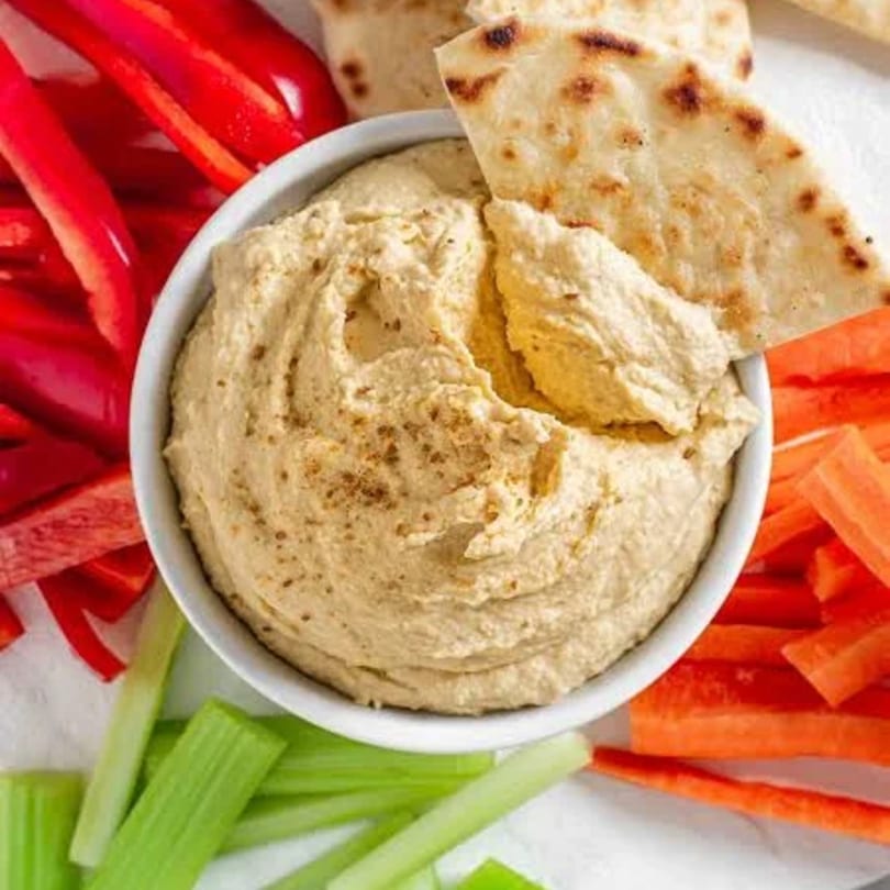 Hummus in a white bowl with pita bread, red bell pepper, carrots, and celery sticks.